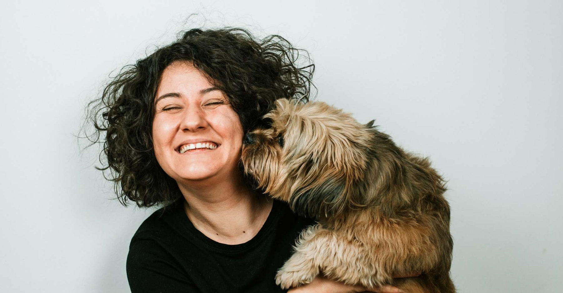 A curly-haired woman holds her dog close while it sniffs her face, hinting at the bond explored in Can Dogs Sense Health Problems in Humans