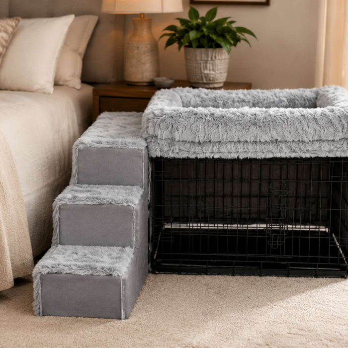 Gray bedside dog bed with plush stairs and a raised cushioned sleeper sits parallel beside a neatly made bed over a black wire crate