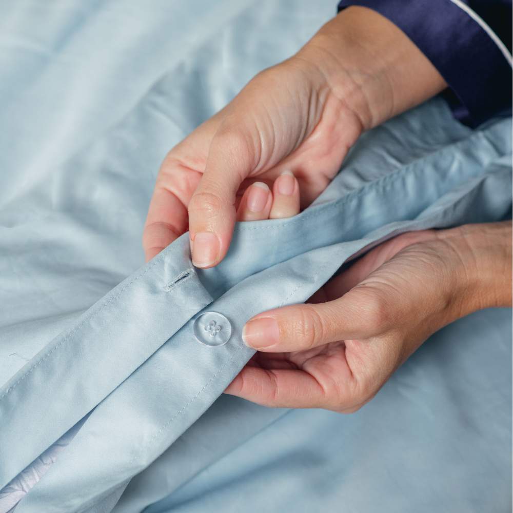 A close-up of hands holding a buttoned area of the bedding, showing the details of the Paw PupSheets™ Hair Resistant, Antimicrobial, & Cooling Duvet Cover and Sheet Set Bundle - Sky Blue