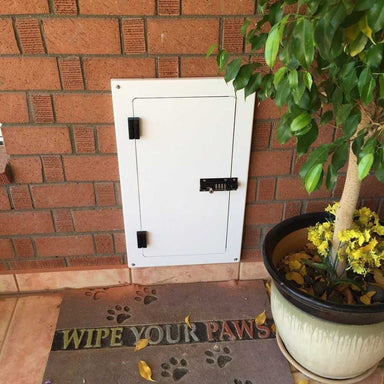 A fully closed Watchdog Security Pet Doors Cover installed in a brick wall with a black combination lock. It's beside a potted plant and a Wipe Your Paws doormat