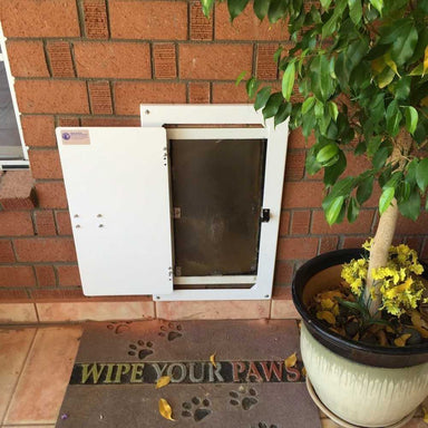 A fully closed Watchdog Security Pet Doors Cover with a black combination lock, installed in a brick wall next to a Wipe Your Paws doormat and a potted plant