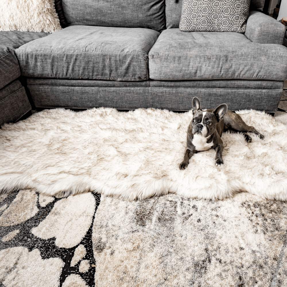 A grey dog lying on a Paw PupRug™ Runner Faux Fur Memory Foam Dog Bed Curve White with Brown Accents in front of a grey sectional couch with decorative pillows