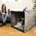 A happy dog sits inside a black wire crate covered with grey fabric, part of the Paw Upgrade Your Dog Crate Kit - Charcoal Grey, while a woman enjoys her coffee nearby