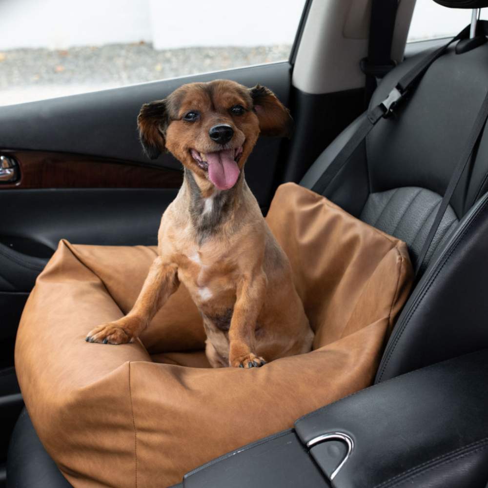 A happy small dog is seated in the Paw PupProtector™ Faux Leather Memory Foam Dog Car Bed - Camel Single Car Seat, placed in the front passenger seat of a car