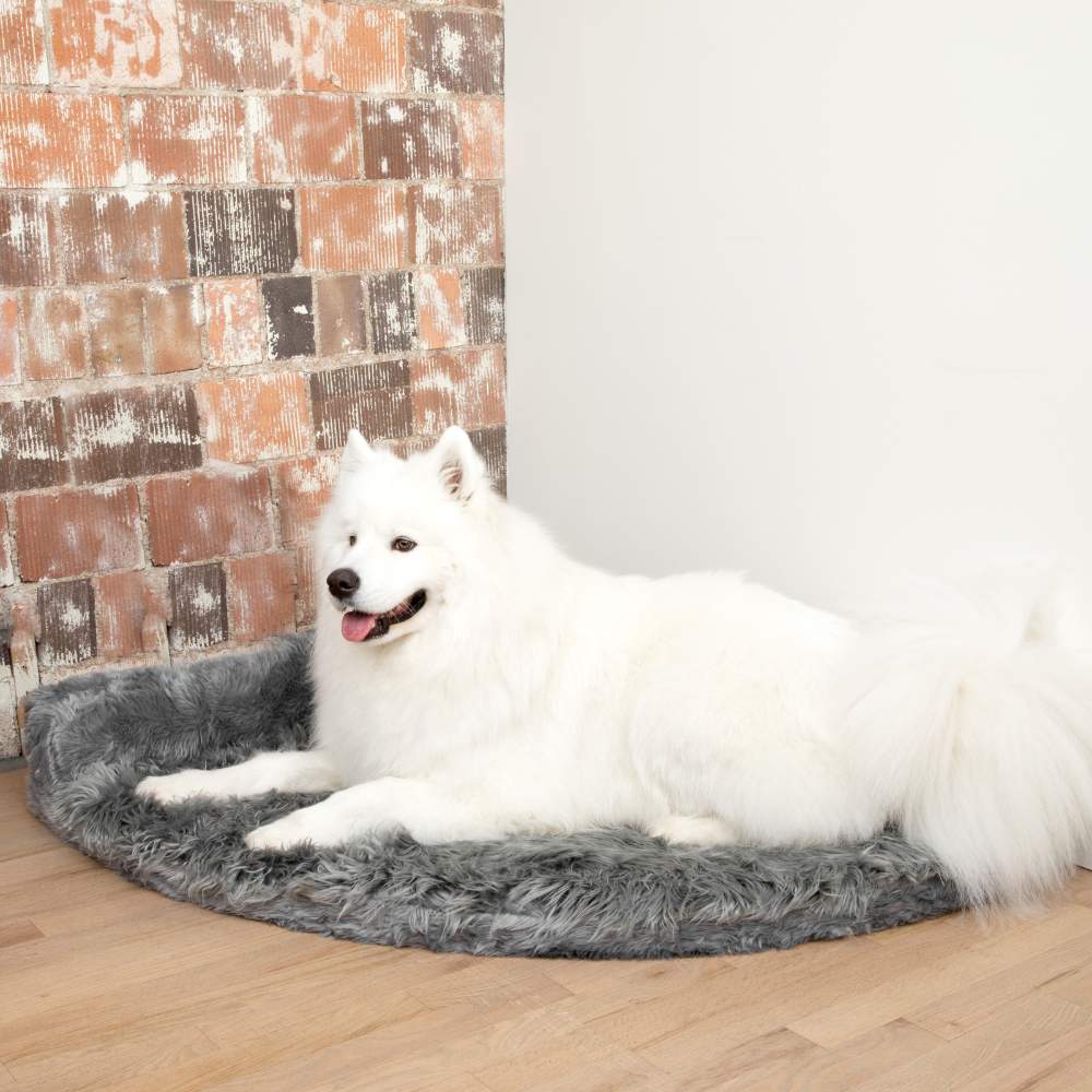 A large fluffy white dog is lying on the Charcoal Grey Paw PupRug™ Memory Foam Corner Dog Bed next to a rustic brick wall