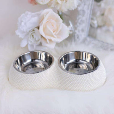 An image shows a Hello Doggie Pearl Dining Bowl with two stainless steel bowls set in a textured, pearl-colored base, surrounded by soft, white decor and flowers in the background