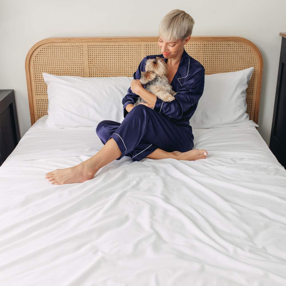A person in navy pajamas sits on a bed holding a small dog, showcasing the Paw PupSheets™ Hair Resistant, Antimicrobial, & Cooling Bed Sheet Set - White