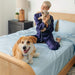 A person in navy pajamas sits on a bed with a large and a small dog, showcasing the Paw PupSheets™ Hair Resistant, Antimicrobial, & Cooling Bed Sheet Set - Sky Blue