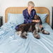 A person in navy pajamas sits on a bed with a large dog, highlighting the Paw PupSheets™ Hair Resistant, Antimicrobial, & Cooling Bed Sheet Set - Sky Blue