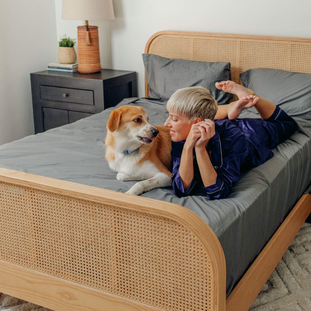 A person lies on a bed with a large dog, showcasing the Paw PupSheets™ Hair Resistant, Antimicrobial, & Cooling Bed Sheet Set - Graphite