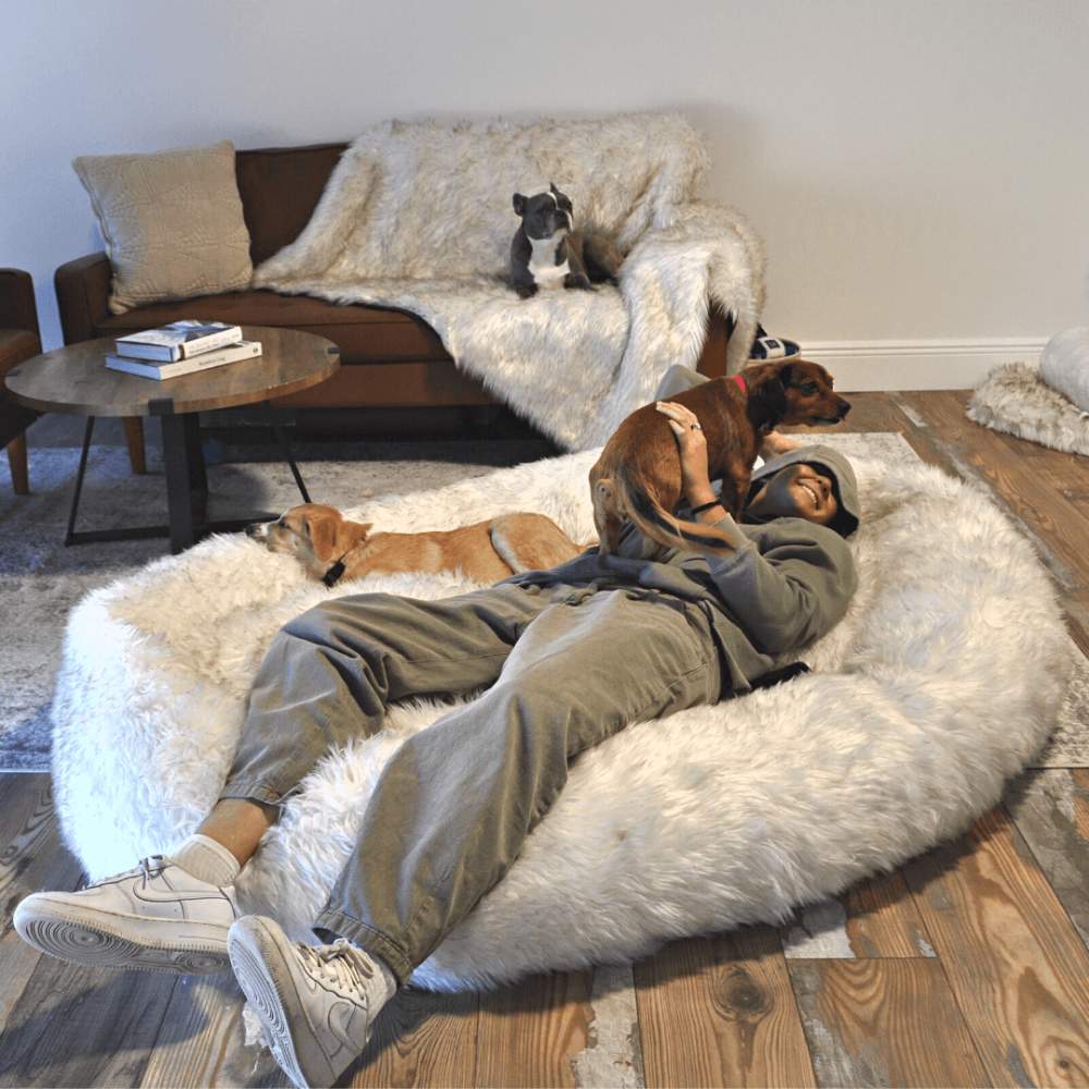 A person relaxes on a White with Brown Accents Paw PupCloud™ Human-Size Faux Fur Memory Foam Dog Bed while two dogs rest beside them