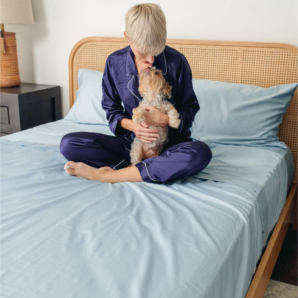 A person sits on a bed holding a small dog with light blue sheets, demonstrating the Paw PupSheets™ Hair Resistant, Antimicrobial, & Cooling Bed Sheet Set - Sky Blue
