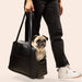 A pug being carried by a person in the Paw PupTote™ 3-in-1 Faux Leather Dog Carrier Bag - Black, with the person’s legs and feet visible