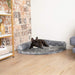 A small black dog with white markings is resting on the Charcoal Grey Paw PupRug™ Memory Foam Corner Dog Bed, situated near a rustic brick wall, a bookshelf, and a blue couch