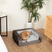 A small brown dog is curled up on the Paw PupChill™ Cooling Bolster Dog Bed, located in a neatly decorated room with a plant