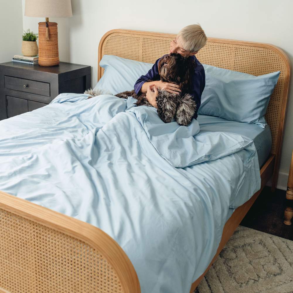 A woman cuddles with her dog on a bed made with the Paw PupSheets™ Hair Resistant, Antimicrobial, & Cooling Duvet Cover and Sham Set - Sky Blue