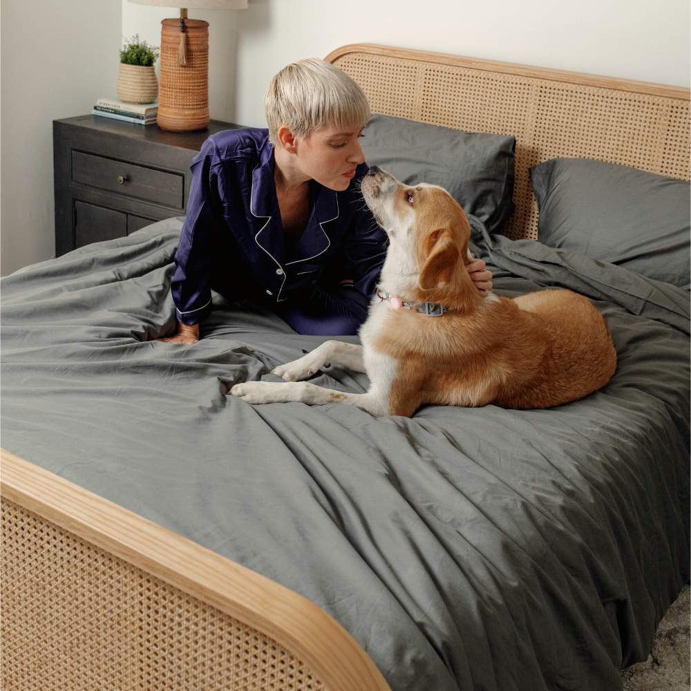A woman in pajamas leaning towards her dog on a bed, both surrounded by the Paw PupSheets™ Hair Resistant, Antimicrobial, & Cooling Duvet Cover and Sheet Set Bundle - Graphite