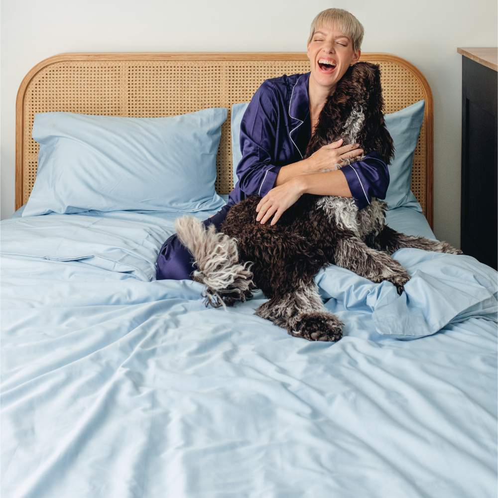 A woman laughing while cuddling her dog on a bed, covered with the Paw PupSheets™ Hair Resistant, Antimicrobial, & Cooling Duvet Cover and Sheet Set Bundle - Sky Blue