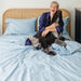 A woman laughing while cuddling her dog on a bed, covered with the Paw PupSheets™ Hair Resistant, Antimicrobial, & Cooling Duvet Cover and Sheet Set Bundle - Sky Blue