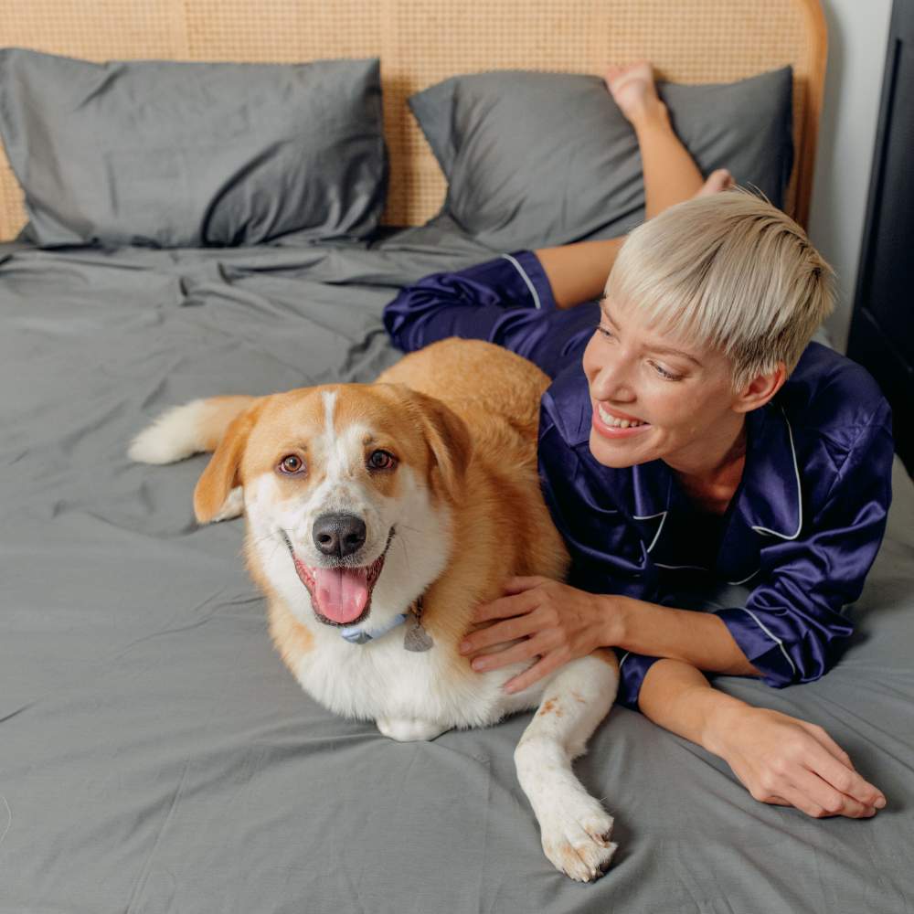 A woman lying beside her dog on a bed, both enjoying the Paw PupSheets™ Hair Resistant, Antimicrobial, & Cooling Duvet Cover and Sheet Set Bundle - Graphite