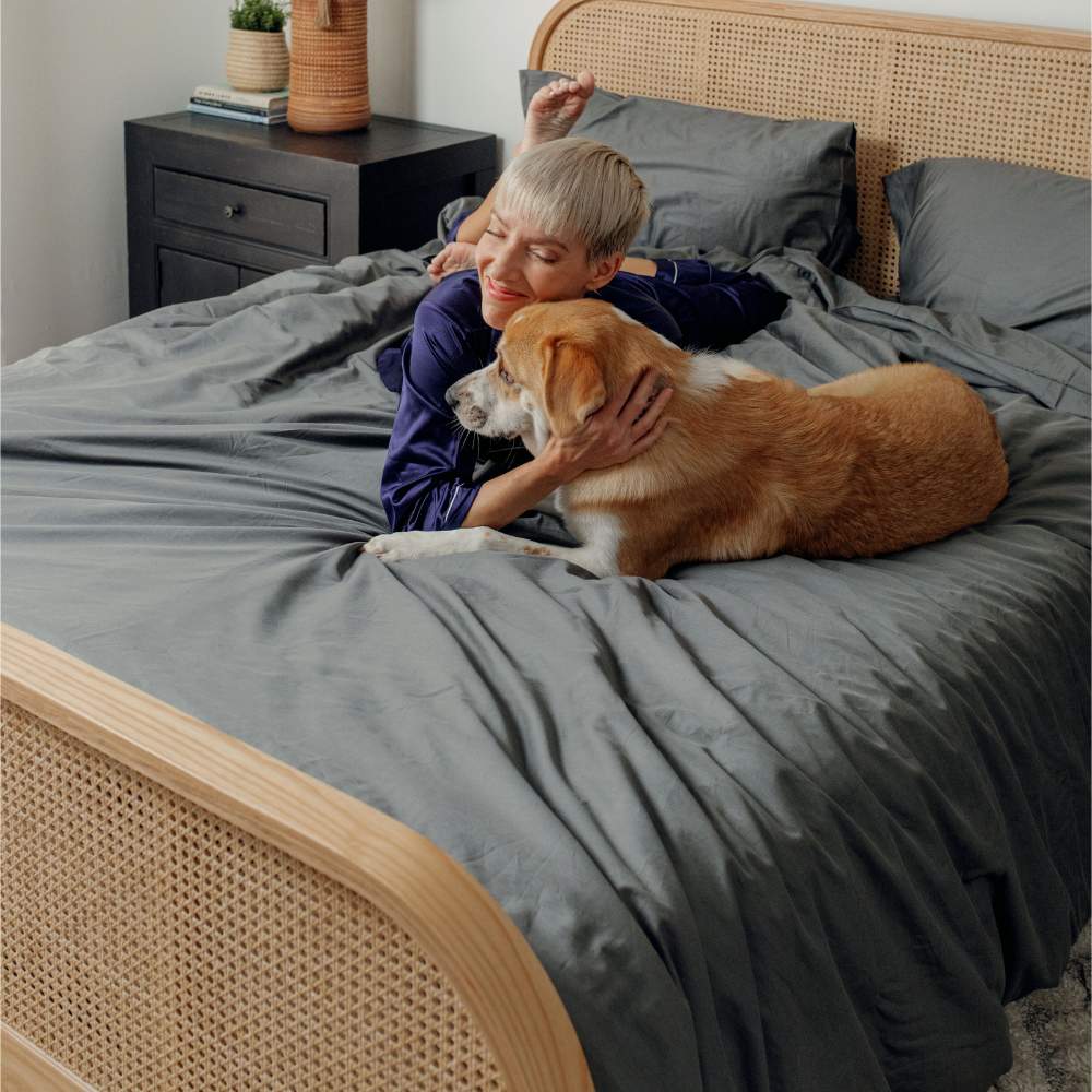 A woman lying on a bed with her dog, both enjoying the comfort of the Paw PupSheets™ Hair Resistant, Antimicrobial, & Cooling Duvet Cover and Sheet Set Bundle - Graphite