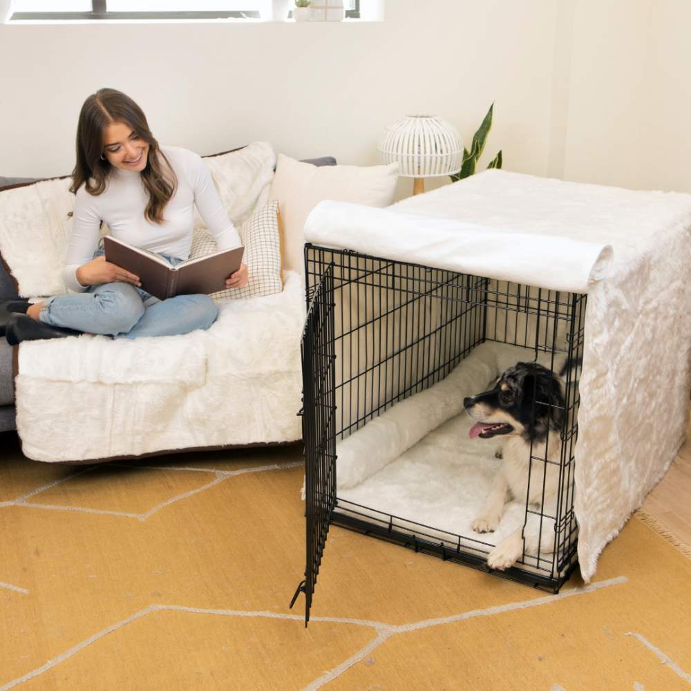 A woman reads a book on the couch while a dog relaxes in a black wire crate with a cozy white interior, part of the Paw Upgrade Your Dog Crate Kit - Polar White