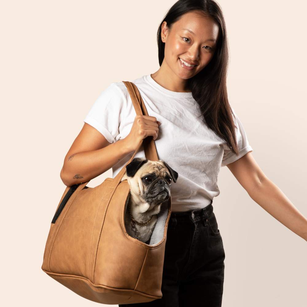 A woman smiling and carrying a pug in the Paw PupTote™ 3-in-1 Faux Leather Dog Carrier Bag - Camel against a plain background