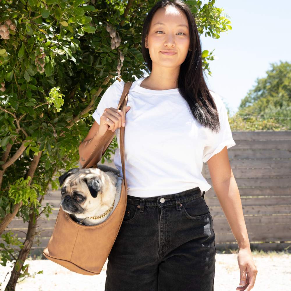 A woman standing outside carrying a pug in the Paw PupTote™ 3-in-1 Faux Leather Dog Carrier Bag - Camel on a sunny day