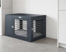 Bowsers Moderno Dog Crate sits in a clean modern room, showing its front and side doors with silver metal bars and a cushioned interior