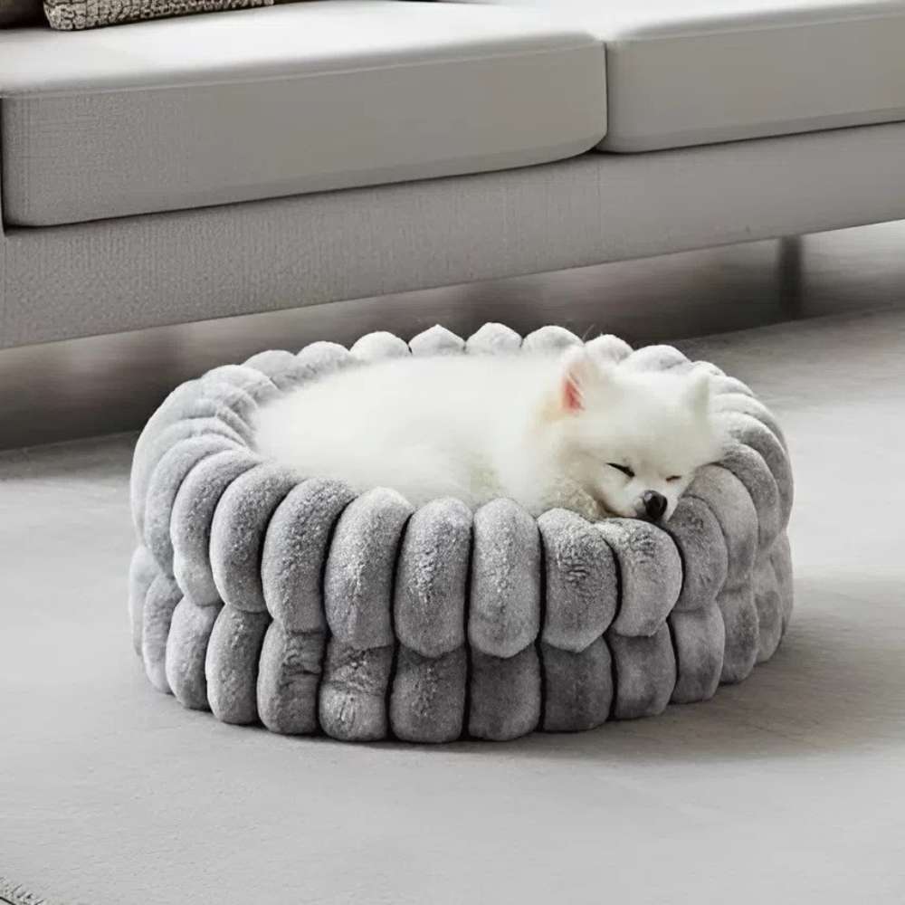 Bowsers The Muffy Dog Bed shows a small white dog sleeping in a soft, gray, round bed with thick padded sides placed in front of a couch