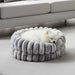 Bowsers The Muffy Dog Bed shows a small white dog sleeping in a soft, gray, round bed with thick padded sides placed in front of a couch