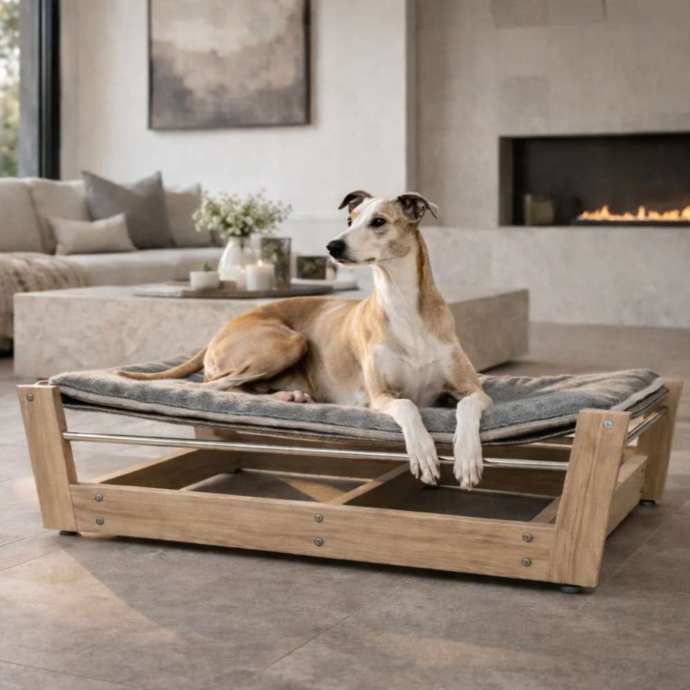 Light-colored dog lounges indoors on a wooden raised bed fitted with Bowsers Yugen Reversible Pad, placed in a cozy living room near a fireplace