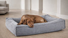 Relaxed brown dog lies curled up on a soft gray Bowsers Divine Futon Dog Bed with thick padded bolsters and a plush sleeping area